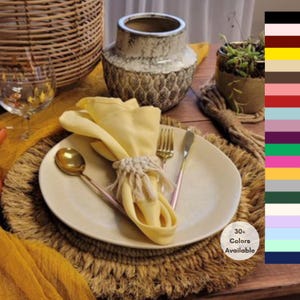 May include: A round, textured jute placemat topped with a cream-colored ceramic plate, a yellow cloth napkin secured by a macrame ring, a gold-toned spoon, a gold-toned fork, and a gold-toned knife. A color chart with 30+ color options is visible.