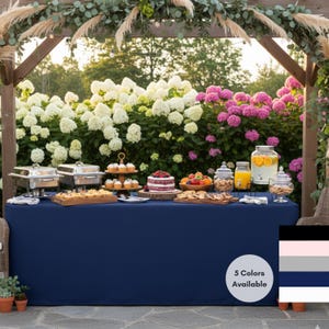 May include: A navy blue rectangular tablecloth covers a table laden with food and drinks. The table is set up outdoors with a wooden arbor and a backdrop of white and pink flowers. Various dishes, cakes, and beverages are displayed.