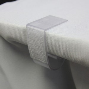 May include: A clear plastic table clip with a hook and a strip of hook and loop fastener. The clip is designed to hold a tablecloth in place.