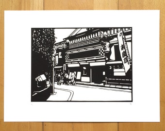 May include: Black and white woodcut print of a street scene in Japan. The print features a traditional Japanese building with a sign that reads "酒場". There are people walking on the street in front of the building.