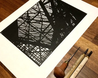 May include: A black and white paper cut art print of a building's structural framework. The print is framed and displayed on a wooden surface with three tools used for paper cutting.