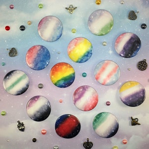 May include: A collection of 14 round pins with different galaxy-themed designs. The pins are arranged in a circle on a light blue background with white speckles. The pins feature various colors, including blue, green, purple, pink, yellow, and white.