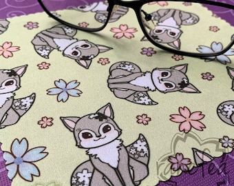 Puede incluir: Un paño de limpieza de microfibra verde claro con un patrón repetido de zorros y flores de dibujos animados. Las gafas negras descansan sobre el paño. Los zorros son grises y blancos con detalles florales, y las flores son rosas y azules.