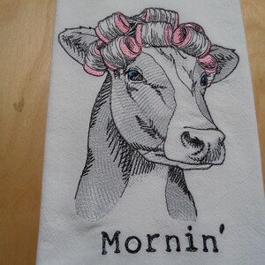 May include: A black and white embroidered illustration of a cow with pink hair rollers, with the text "Mornin'" below the image.