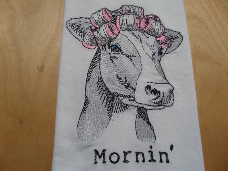 May include: A black and white embroidered illustration of a cow with pink hair rollers, with the text "Mornin'" below the image.