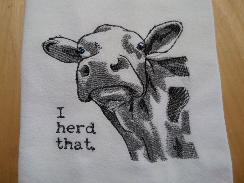 May include: A white kitchen towel with a black and white embroidered image of a cow's head with the text "I herd that."