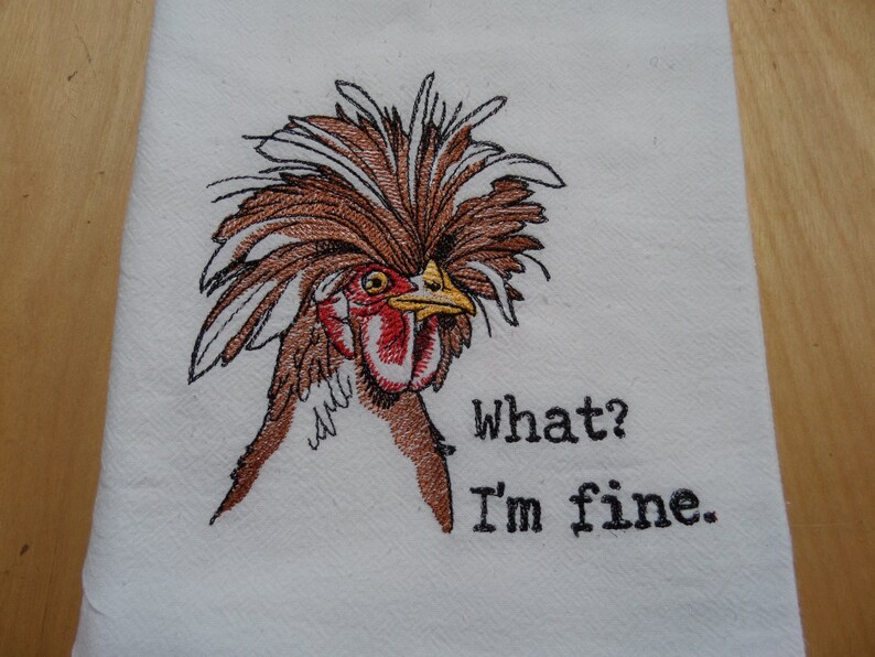 May include: Embroidered kitchen towel featuring a brown and white chicken with a large, fluffy crest. The chicken is looking to the right and the text "What? I'm fine." is embroidered below the chicken.