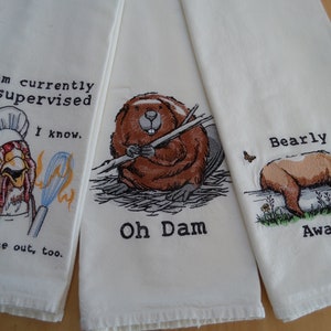 May include: Three white kitchen towels with embroidered images and text. The first towel features a cartoon chicken wearing a chef's hat and holding a whisk, with the text "I am currently unsupervised I know. freaks me out, too." The second towel features a cartoon beaver holding a branch with the text "Oh Dam." The third towel features a cartoon bear lying down with the text "Bearly Awake."
