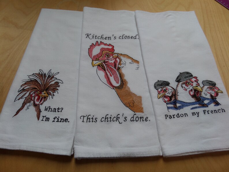 May include: Three white kitchen towels with embroidered chickens and humorous sayings. The first towel features a chicken with a mohawk and the text "What? I'm fine." The second towel features a rooster with the text "Kitchen's closed. This chick's done." The third towel features three chickens wearing berets and the text "Pardon my French."
