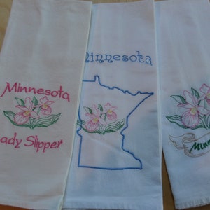 May include: Three white tea towels with embroidered designs. The first towel features a pink and green lady slipper flower with the text "Minnesota Lady Slipper". The second towel features a blue outline of the state of Minnesota with a pink and green lady slipper flower and the text "Minnesota". The third towel features a pink and green lady slipper flower with a brown ribbon that says "Minnesota".