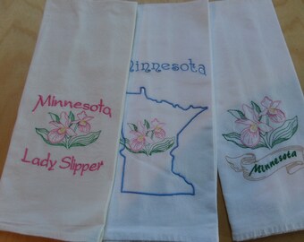 May include: Three white tea towels with embroidered designs. The first towel features a pink and green lady slipper flower with the text "Minnesota Lady Slipper". The second towel features a blue outline of the state of Minnesota with a pink and green lady slipper flower and the text "Minnesota". The third towel features a pink and green lady slipper flower with a brown ribbon that says "Minnesota".