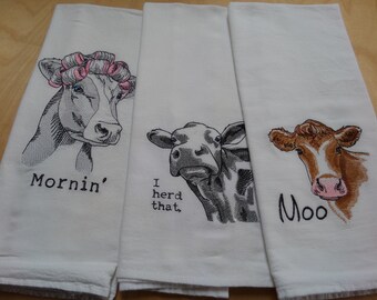 May include: Three white kitchen towels with embroidered cow designs. The first towel has a cow with pink hair rollers and the text "Mornin'" below. The second towel has a cow with the text "I herd that." below. The third towel has a brown cow with the text "Moo" below.