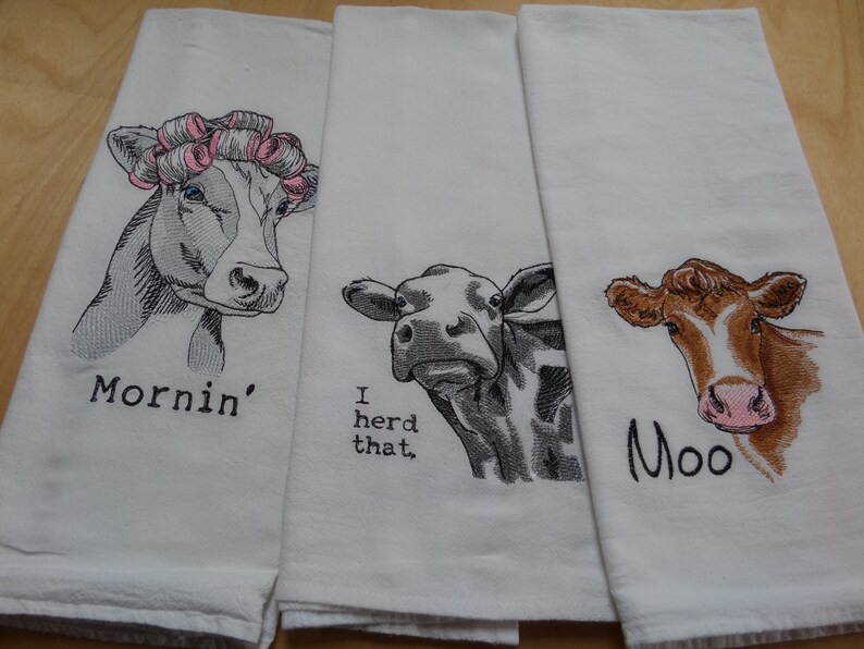 May include: Three white kitchen towels with embroidered cow designs. The first towel has a cow with pink hair rollers and the text "Mornin'" below. The second towel has a cow with the text "I herd that." below. The third towel has a brown cow with the text "Moo" below.