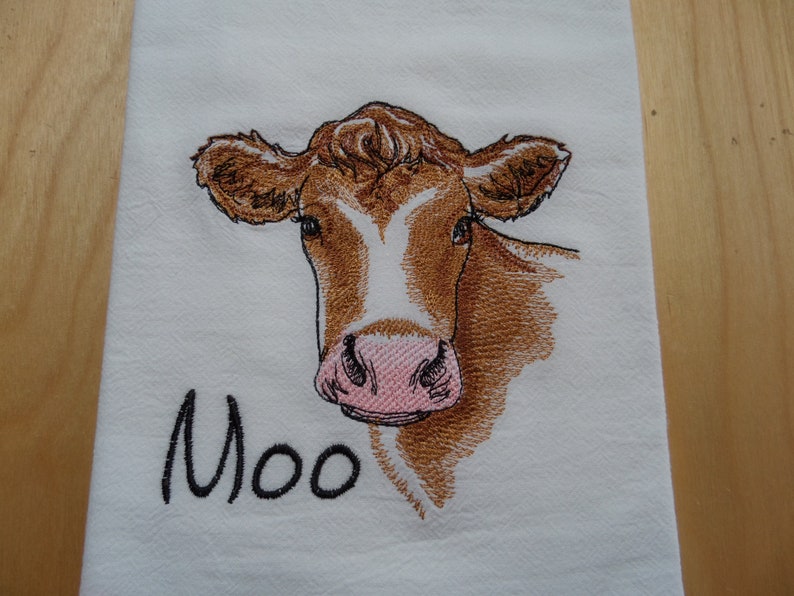 May include: Embroidered kitchen towel featuring a brown and white cow with a pink nose. The towel has the word "Moo" embroidered below the cow.