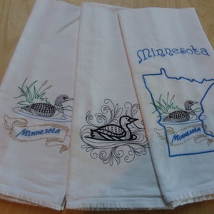 May include: Three white kitchen towels featuring embroidered designs. One towel shows a loon on water with the text Minnesota on a ribbon. Another displays a loon surrounded by swirling patterns. The third features a loon inside a blue outline of Minnesota.