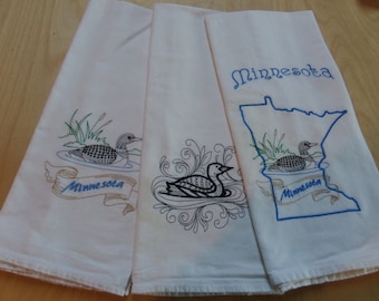 May include: Three white kitchen towels featuring embroidered designs. One towel shows a loon on water with the text Minnesota on a ribbon. Another displays a loon surrounded by swirling patterns. The third features a loon inside a blue outline of Minnesota.