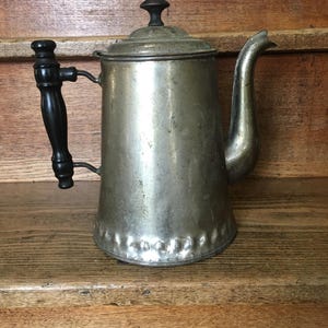 May include: An antique silver-toned metal coffee pot with a black wooden handle. The pot has a curved spout and a lid with a small black knob. The pot is sitting on a wooden surface.