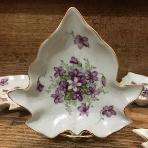 May include: A white, leaf-shaped dish with a gold rim and a floral design featuring purple and white flowers. The dish is part of a set, with other similar dishes visible in the background. The design includes green leaves.