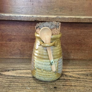 May include: A handmade ceramic jar with a cork stopper and a small wooden spoon. The jar is a mix of tan, brown, and gray colors. The spoon is attached to the jar with a small loop. The jar is sitting on a wooden surface.