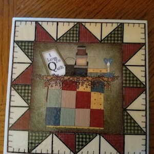 May include: A white ceramic tile with a patchwork quilt design. The tile features a framed sign that reads "I Love To Quilt." The tile is surrounded by a border of red, green, and white triangles.