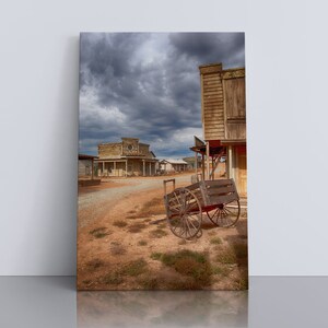 May include: A weathered wooden wagon sits in the middle of a deserted Western town street. The buildings are made of wood and have a rustic look. The sky is overcast with dark clouds.