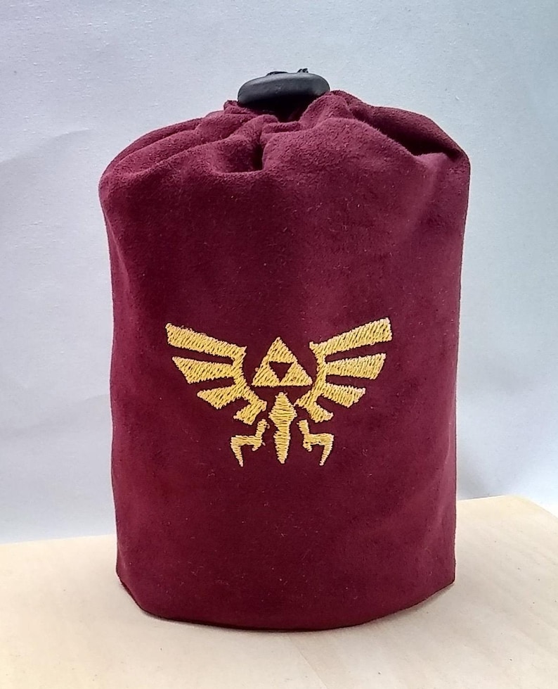 Pu&ograve; includere: Una borsa a cordoncino in velluto bordeaux con un simbolo Triforce ricamato in oro.