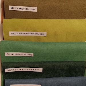 Pu&ograve; includere: Una collezione di campioni di tessuto in vari colori e trame, tra cui beige, oro, kiwi, oliva, verde neon, verde, verde scuro, blu chiaro e blu. I campioni sono etichettati con i rispettivi nomi e materiali, come "Beige Alova Knit" e "Olive Microsuede".