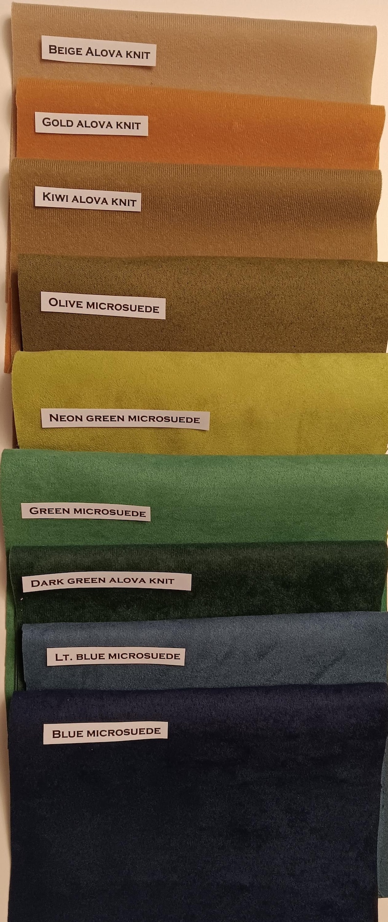 Pu&ograve; includere: Una collezione di campioni di tessuto in vari colori e trame, tra cui beige, oro, kiwi, oliva, verde neon, verde, verde scuro, blu chiaro e blu. I campioni sono etichettati con i rispettivi nomi e materiali, come "Beige Alova Knit" e "Olive Microsuede".