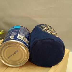 Pu&ograve; includere: Una pochette in tessuto blu con un ricamo dorato. La pochette &egrave; appoggiata su una superficie di legno accanto a una lattina argentata con un'etichetta blu e bianca.