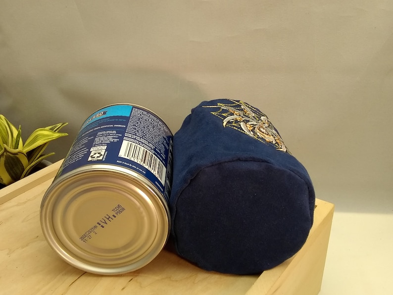 Pu&ograve; includere: Una pochette in tessuto blu con un ricamo dorato. La pochette &egrave; appoggiata su una superficie di legno accanto a una lattina argentata con un'etichetta blu e bianca.