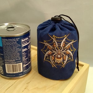 Pu&ograve; includere: Una borsa a cordoncino blu con un ragno bianco e una ragnatela ricamate sul davanti. La borsa &egrave; appoggiata su una superficie di legno accanto a una lattina di zuppa.
