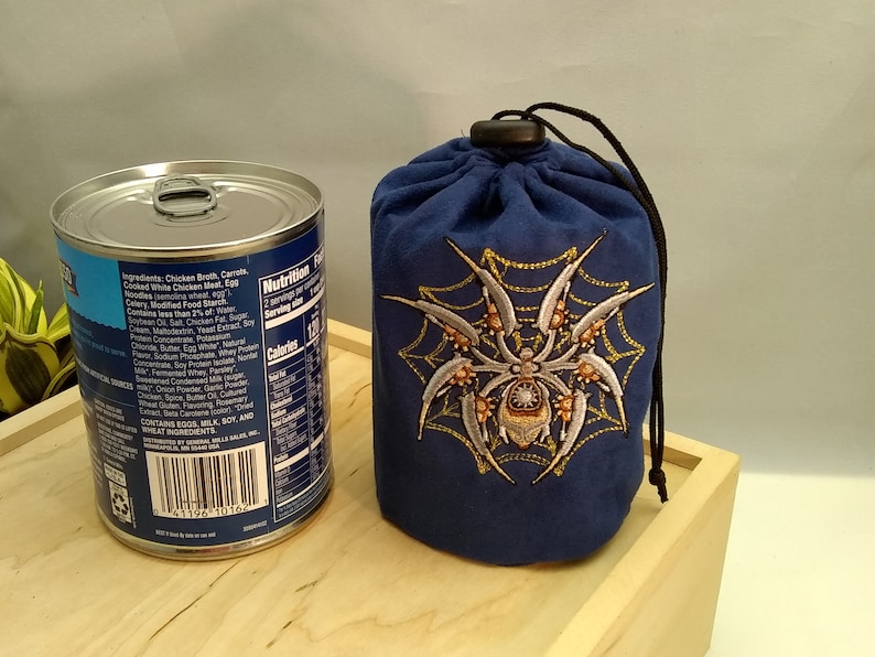 Pu&ograve; includere: Una borsa a cordoncino blu con un ragno bianco e una ragnatela ricamate sul davanti. La borsa &egrave; appoggiata su una superficie di legno accanto a una lattina di zuppa.