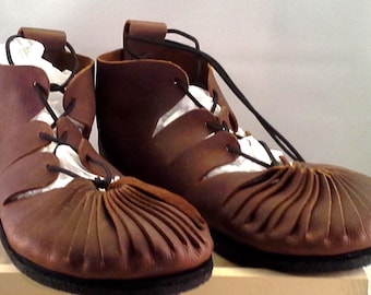 May include: A pair of brown leather shoes featuring a gathered, pleated toe design and black laces that thread through side cutouts. The shoes have a high-top ankle profile with a pull tab at the back and a solid black sole, showcasing a distinct construction.