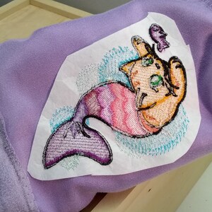Pu&ograve; includere: Un tessuto viola con un'immagine ricamata di un gatto con una coda di sirena. Il gatto &egrave; arancione e la coda &egrave; viola e rosa. Il gatto tiene un piccolo pesce viola in bocca.
