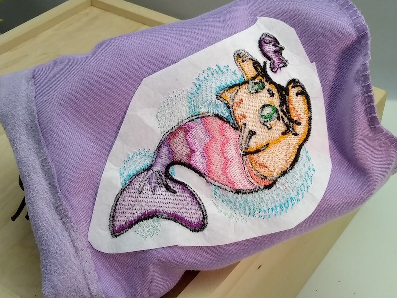 Pu&ograve; includere: Un tessuto viola con un'immagine ricamata di un gatto con una coda di sirena. Il gatto &egrave; arancione e la coda &egrave; viola e rosa. Il gatto tiene un piccolo pesce viola in bocca.