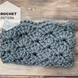 May include: A gray crocheted headband with a textured pattern. The headband is on a wooden surface. CROCHET PATTERN is written in white text on a white hexagon.