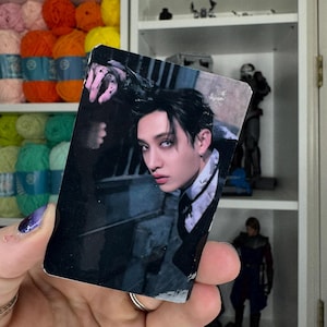 May include: A rectangular trading card featuring a person with dark hair and pale skin, wearing a dark suit and tie. The card has a dark background and the person is looking at the viewer. The card is held in a hand with painted nails.