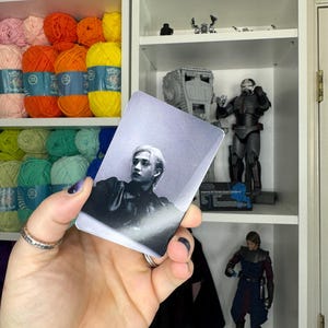 May include: A grayscale photograph of a person on a rectangular card. The card is held in front of a shelf with yarn and Star Wars figurines. The person in the photo has light-colored hair and is wearing a dark jacket.