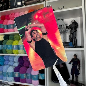 Puede incluir: Una pequeña imagen rectangular de una persona con una camiseta sin mangas negra, sosteniendo un arma, con un fondo rojo y naranja. La imagen está siendo sostenida por unas pinzas plateadas. Estantes en el fondo llenos de hilo y artículos de Star Wars.