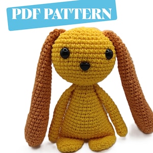 May include: A handmade crochet bunny in shades of yellow and brown. The bunny has long ears, black safety eyes, and a small black nose. The text "PDF PATTERN" is in a blue rectangle above the bunny. This item is a pattern for a crochet project.