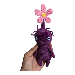 May include: A purple crocheted plush toy with a pink flower on top. The toy has a round body and two arms. It has two white eyes with black pupils.