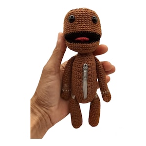 May include: Brown crocheted plush toy with a zipper down the front. The toy has a large mouth with a red tongue sticking out and black eyes.
