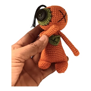 Puede incluir: Muñeca naranja de ganchillo con un ojo de botón, detalles verdes y detalles cosidos en negro. La muñeca hecha a mano tiene un diseño único, con un ojo de botón y un detalle en forma de hoja en el pecho. La muñeca se sostiene en una mano.