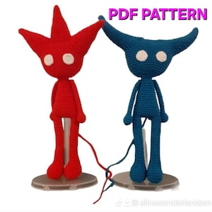 Puede incluir: Patrón PDF para tejer dos figuras amigurumi. Una figura es roja con una cabeza en forma de estrella y la otra es azul con cuernos. Ambas figuras tienen ojos blancos y están de pie sobre bases transparentes.