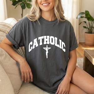 Puede incluir: Camiseta gris oscuro con la palabra "CATHOLIC" en blanco arqueada en la parte superior y un crucifijo blanco debajo. La camiseta tiene mangas enrolladas y un ajuste relajado. La camiseta está hecha de un material suave.