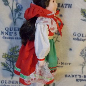 May include: A porcelain doll wearing a red, white, and green dress with a white lace trim. The doll has brown hair and is wearing white shoes.