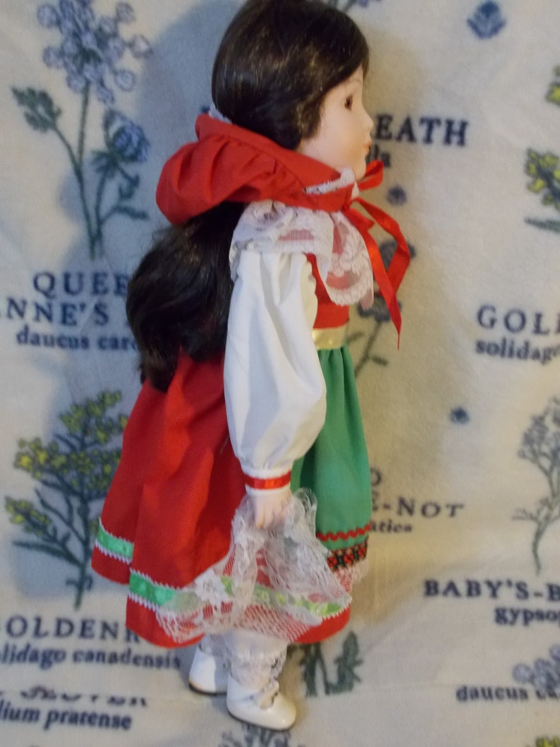 May include: A porcelain doll wearing a red, white, and green dress with a white lace trim. The doll has brown hair and is wearing white shoes.
