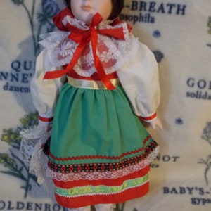May include: A porcelain doll wearing a white blouse with a red and white lace collar, a red bow, and a green skirt with red, white, and black trim. The doll has brown hair and brown eyes.