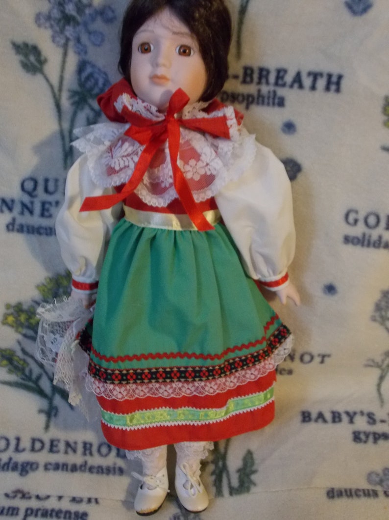 May include: A porcelain doll wearing a white blouse with a red and white lace collar, a red bow, and a green skirt with red, white, and black trim. The doll has brown hair and brown eyes.