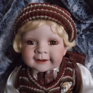 Peut inclure: Une poupée en porcelaine aux cheveux blonds portant un pull rayé marron et blanc et un bonnet en tricot marron. La poupée sourit et a les yeux marrons.